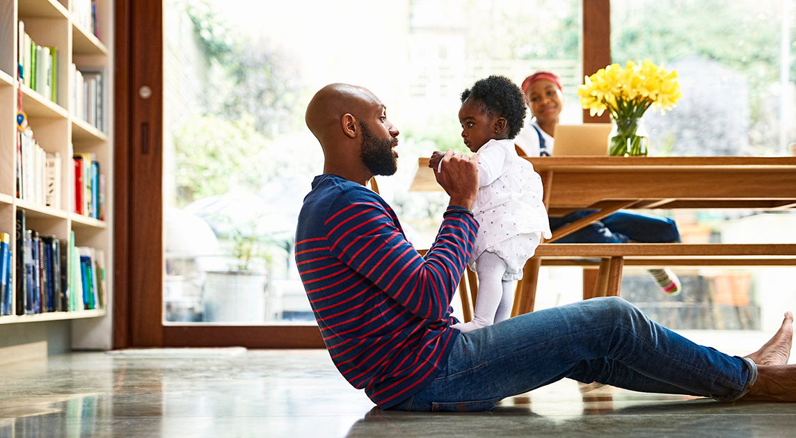 A Covered California member who has saved money on health insurance through   the American Rescue Plan plays in his living room with is baby and wife.